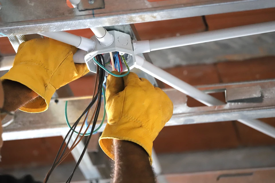 Électricien en combinaison bleue et gants blancs utilisant un multimètre jaune pour tester un tableau électrique industriel